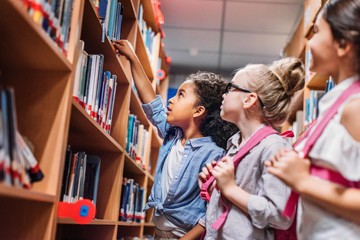Three young girls looking at books on shelf in library.
